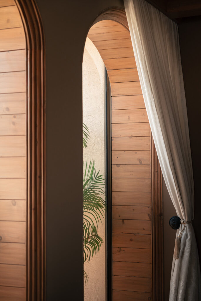 Close view of a timber-lined arched window with a pale curtain and greenery beyond at Still-Wood.