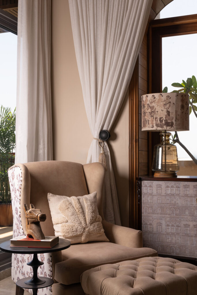 A tufted armchair and matching ottoman beside a patterned table lamp, cabinet and pale curtain at Still-Wood.