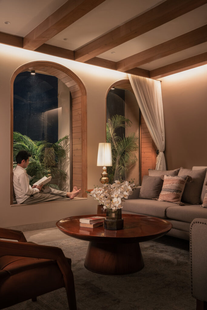 The Still-Wood living room at night, with a flower-topped timber coffee table, cream sofas and greenery beyond the arched windows.